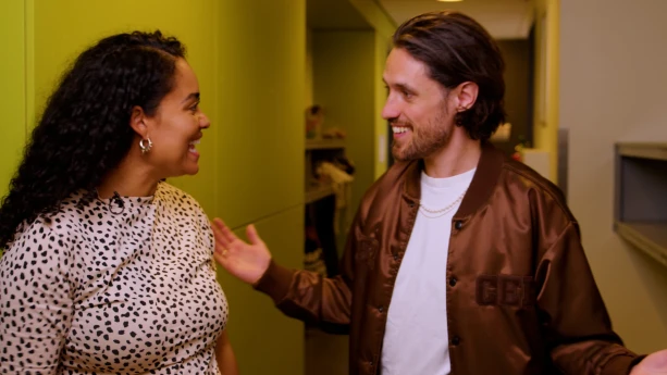 Een foto van presentator Ciana. Ze praat enthousiast met gast Jasper. Ze staan in een smalle gang.