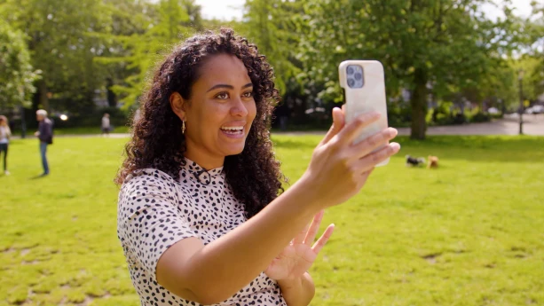 Een foto van presentator Ciana in een park. Ze praat en maakt een filmpje van zichzelf met haar telefoon. Achter haar zijn wat mensen en honden.....