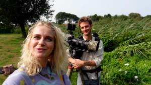 Een foto van Janouk en Bram in een grasveld met rechts naast hen zichtbare struiken en achter hen een boom. Bram heeft een grote fotocamera op een statief vast.