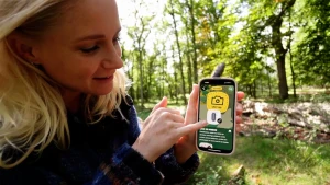 Een foto van presentator Janouk in het bos. Ze wijst op het scherm van de mobiele telefoon in haar hand, waarop een pissebed te zien is.