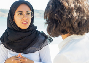 Twee vrouwen in gesprek, een van hen draagt een zwarte hoofddoek en een bril.