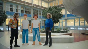 Een foto van de hal van het Amsterdam UMC met daarin een presentator en wetenschapper Baudewijntje Kreukels. Tussen hen in staan twee getekende kartonnen figuren van een jongen en een meisje in een wit t-shirt en een spijkerbroek. 