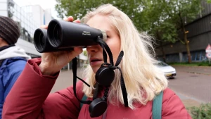 Een foto van presentator Janouk die in haar rechterhand een verrekijker heeft en daardoor kijkt. Janouk staat in het midden van een straat.