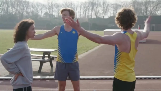 Een foto van een presentator en twee mensen in sportkleding op een sintelbaan. De presentator staat links, de twee mensen aan de rechterkant staan met hun armen gespreid. Een sintelbaan is de loopbaan die om een atletiekbaan heen loopt. 