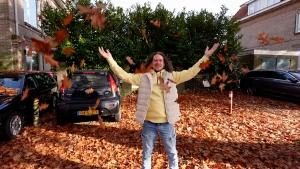 Een foto van een presentator die op een weg in een woonwijk staat die vol ligt met herfstbladeren. De presentator gooit bladeren de lucht in en de bladeren zweven op de foto naar beneden. De bomen waarvan de bladeren afkomen, zijn niet zichtbaar.