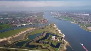 Een foto van de Waal rivier, slot Loevenstein is in beeld samen met verschillende dorpen en een stad. In de rivier vaart er een vrachtschip.