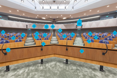 Foto van de vergaderzaal van de Tweede Kamer. Er staan houten bankjes en blauwe zetels. Vooraan staan microfoons. Verdeeld over de foto zijn blauwe icoontjes geplaatst.