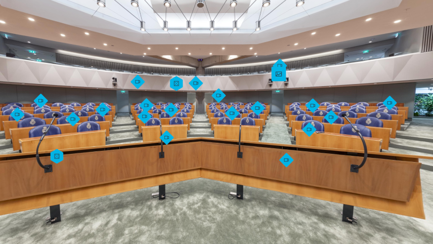 Foto van de vergaderzaal van de Tweede Kamer. Er staan houten bankjes en blauwe zetels. Vooraan staan microfoons. Verdeeld over de foto zijn blauwe icoontjes geplaatst.