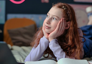 Meisje met rood haar ligt op bed en kijkt dromerig naar buiten met hoofd rustend in haar handen.