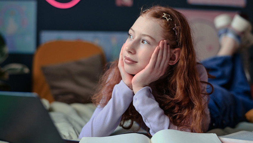 Meisje met rood haar ligt op bed en kijkt dromerig naar buiten met hoofd rustend in haar handen.