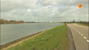 Foto van een dijk met gras met aan de linkerkant een brede rivier en aan de rechterkant een geasfalteerde weg.