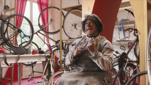 Een foto van Piet die met een werkschort aan in een ruimte zit vol met kapotte fietsen. Piet heeft gereedschap in zijn hand.