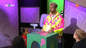 Foto van presentator in fluorescerend jack achter desk met kinderen om hem heen.