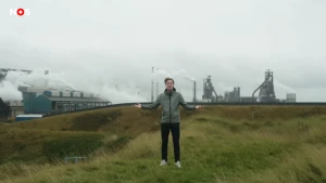 Foto van presentator Dennis die in een veld naast fabrieken staat. De fabrieken stoten grote witte wolken uit.