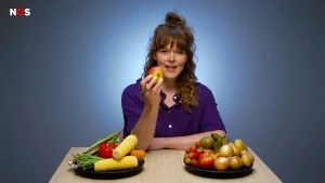 Een foto van presentatrice Jeanine. Voor haar op tafel staan twee schalen. Eén schaal met maïs, paprika en bonen. Een andere schaal met appels, aardbeien en uien. Ze houdt een appel omhoog.