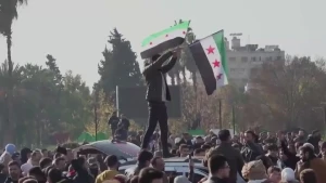   Een foto van een man staat op een auto in een grote menigte, hij zwaait met twee grote vlaggen van de Syrische revolutie.