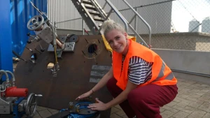 Foto van presentator Janouk met oranje veiligheidshesje die objecten laat zien met metalen platen en draaiwielen die gebruikt worden voor scheepsberging.