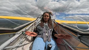 Foto van presentator Eva die zit in de glazen cockpit van een zweefvliegtuig. Door het glas zie je het land op grote afstand. Eva legt iets uit met haar handen.