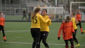 Op deze afbeelding zijn meerdere kinderen te zien die voetballen. Het ene team draagt geel, en het andere oranje. Twee van het gele team geven elkaar een high five en zijn blij.