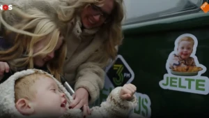 Foto van kind van vier jaar oud in kinderwagen met moeder en zus die over hem heen buigen. Ze kijken allemaal blij naar een foto van Jelte geplakt op de bus van 3FM Serious Request.