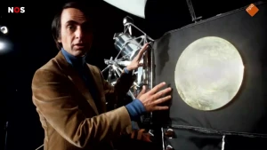 Foto van astronoom Carl Sagan naast ruimtesonde Voyager 1, een grote zwarte kubusachtige vorm met uitsteeksels.