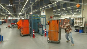 Foto van presentator Bart die een kar met een grote oranje postzak duwt in een grote industriële hal.