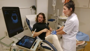 Foto van presentator Pascal in een behandelingsstoel. Een dokter met witte jas wrijft met een echo-apparaat over zijn buik. Ze kijken allebei naar een scherm waar vage contouren op verschijnen.