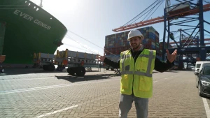 Foto van presentator Nizar die aan de kade staat van een haven. Achter hem een deel van een heel groot vrachtschip en grote hijskranen.