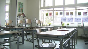 Op deze afbeelding is een leeg klaslokaal te zien. Enkele stoelen staan op tafel en een paar niet. Het daglicht valt binnen via de grote ramen en er hangen knutselwerkjes aan het raam.