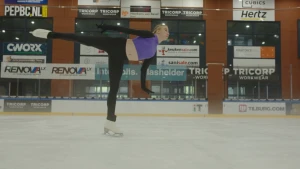 Een foto van kunstschaatser Lindsay van Zundert op een ijsbaan binnen. Ze draagt witte kunstschaatsen en schaatst op één been met haar andere been in de lucht, haar armen sierlijk langs haar lichaam.