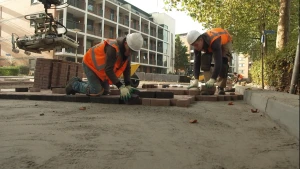 Een foto van twee stratenmakers aan het werk. Ze dragen oranje hesjes en witte helmen. Ze leggen de stenen op een vlakke ondergrond van zand. Achter hen staat een grijper die een hele stapel stenen oppakt.