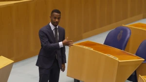 Een foto van Tweede Kamerlid Habtamu de Hoop. Hij staat vooraan in de Tweede Kamer en draagt een net pak. Hij wijst naar de zitplekken van de Tweede Kamerleden, dit zijn blauwe leren stoelen met lange houten bureaus ervoor.