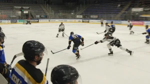 Een foto van een schaatsbaan binnen. Op de voorgrond twee ijshockeyers, eentje met een blauw shirt en eentje met een wit shirt. Die in het witte shirt probeert de puck af te pakken van degene in het blauwe shirt door zijn stick naar voren te steken.