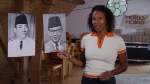 Presentatrice Astrid staat naast een waslijn. Hierop hangen zwart-wit portretten van Soekarno en Hatta.