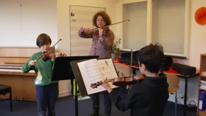 Foto van een muziekstudio met volwassen docent en twee kinderen die viool spelen. Er staat een lessenaar met bladmuziek erop.