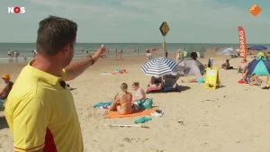Foto van man van de reddingsbrigade die wijst naar een bord op het strand. Er lopen en zitten mensen op het strand.