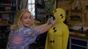 Foto van presentatrice die in de camera kijkt terwijl ze een kettlebell aan een ketting tegen het gezicht van een in het geel geklede crashtestdummie houdt.