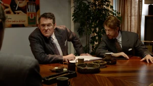 Foto van in pak geklede heren aan donkere houten tafel die samenzweederig naar elkaar toe leunen.