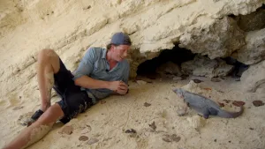 Foto van presentator Freek Vonk die op het strand ligt en naar een leguaan kijkt.