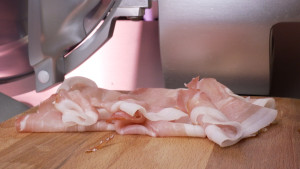 Een foto van dungesneden rauwe plakjes ham die op een snijplank liggen naast een snijmachine.