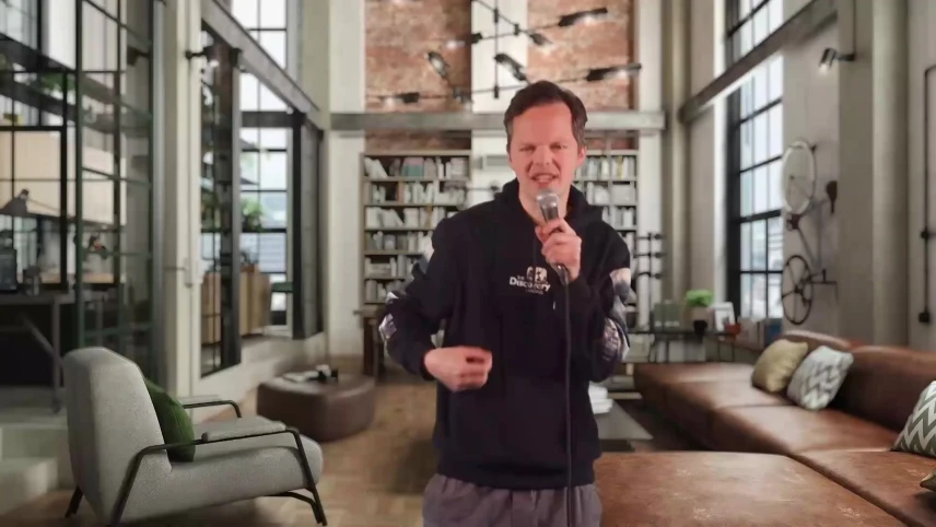 Een foto van presentator Sander Zwiep met een hoodie aan en in zijn hand ene microfoon, op de achtergrond een modern ingerichte huiskamer.