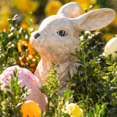 Een foto van een beeldje van de paashaas die tussen de bosjes en de paaseieren in de zon staat.