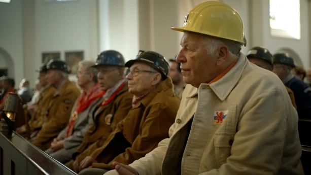 Een foto van oude mensen met mijnwerkershelmen op een rijtje in een bankje, ze lijken ergens naar te kijken/luisteren.