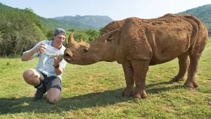 Een foto van Freek Vonk die knielend water geeft aan een grote neushoorn waar hij naast staat.