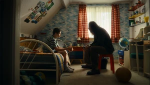 Een foto van een kinderkamer. Links in beeld staat een bed. Daarop zit een jongen. Rechts in beeld zit een man op een plastic kruk. Ze kijken elkaar aan.