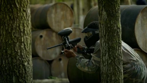 Foto van een persoon met helm en paintballgeweer die hurkt tussen de bomen.