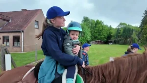 Een afbeelding van twee kinderen op een paard. Ze rijden op straat langs een huis.
