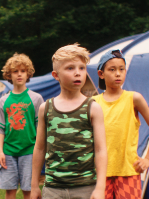 Een foto van kleuters op een cmaping, op de achtergrond zie je een tent en tussen hen in ligt een stapel takken.