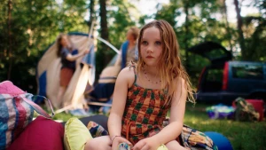 Een foto van een kleutermeisje op een kleed tussen de campingspullen, op de achtergrond zetten twee volwassenen een tent op.