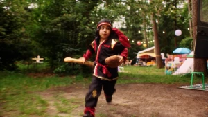 Een foto van een jongetje dat verkleed als Ninja over een camping rent, hij heeft een rood met zwart pak aan en een stokbrood in zn hand.